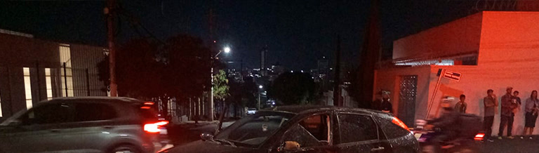 Carro escuro com vidro quebrado e curiosos na calçada após o acidente no Marciano Brandão, à noite