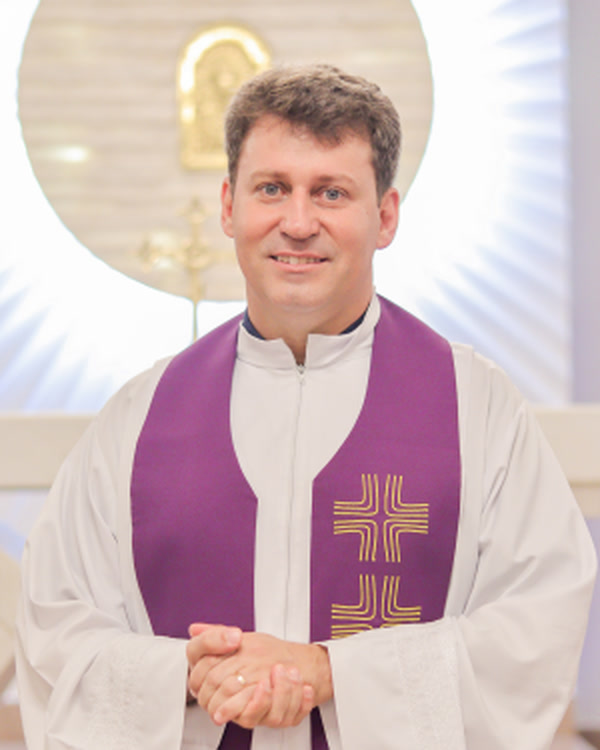 Padre José Luís sorri de túnica branca e estola roxa, com as mãos juntas, diante do altar