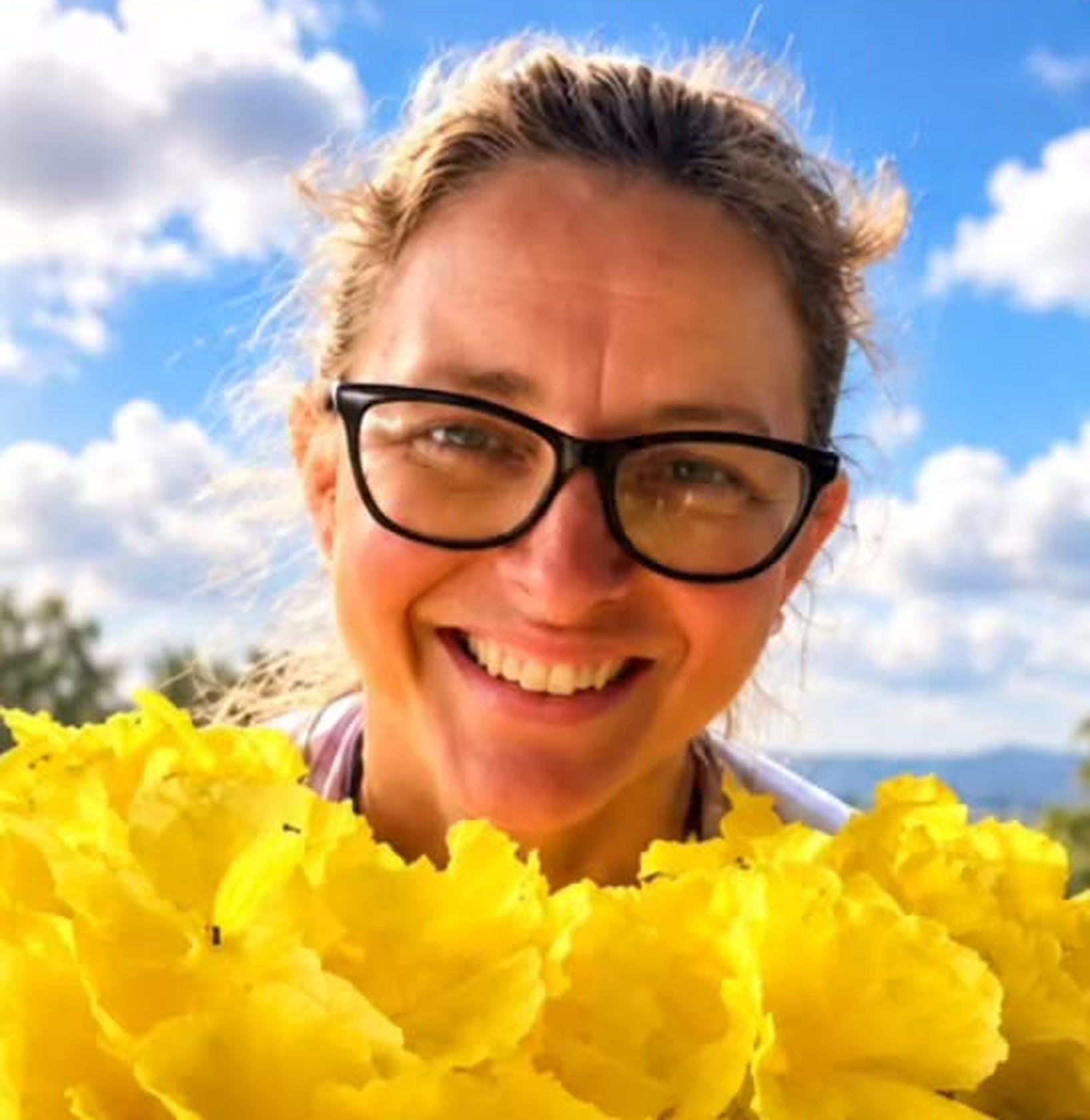 Rosa Maria dos Reis Amaral, a Tia Rosa, sorrindo, de óculos, atrás de um buquê de flores amarelas