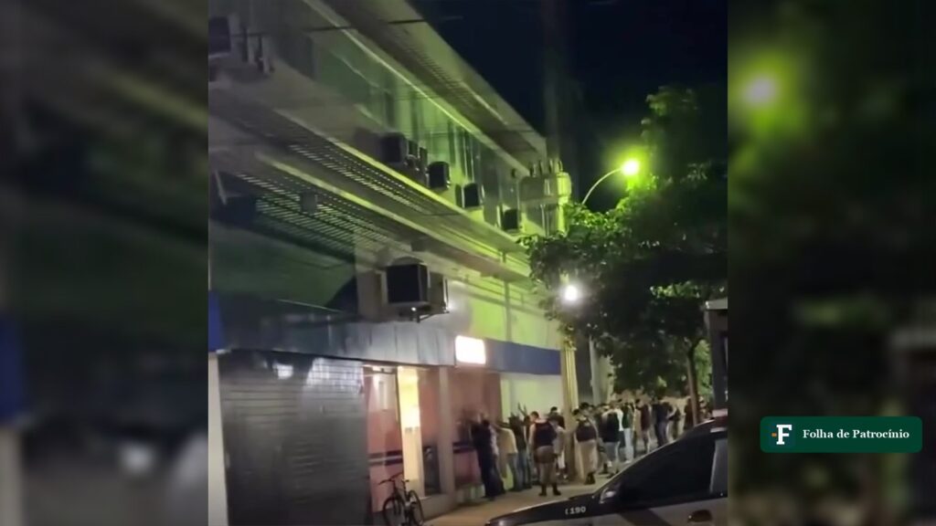Abordagem da PM em João Pinheiro: viatura e grupo encostado na parede ao lado da Caixa, à noite