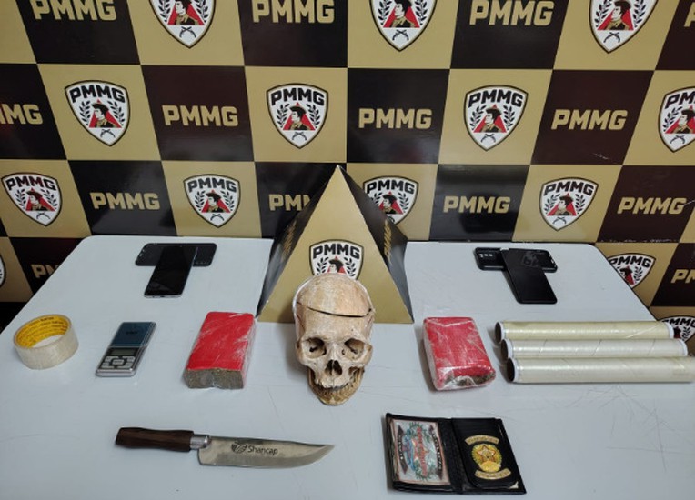 Material da apreensão em Coromandel exposto sobre a mesa: drogas, balança, objeto semelhante a crânio e carteira com brasão da Polícia Civil