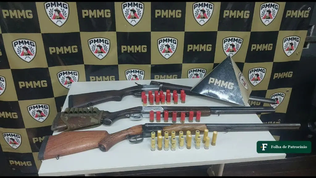 Três espingardas e munições apreendidas pela PM em Salitre de Minas, sobre mesa diante do painel da PMMG