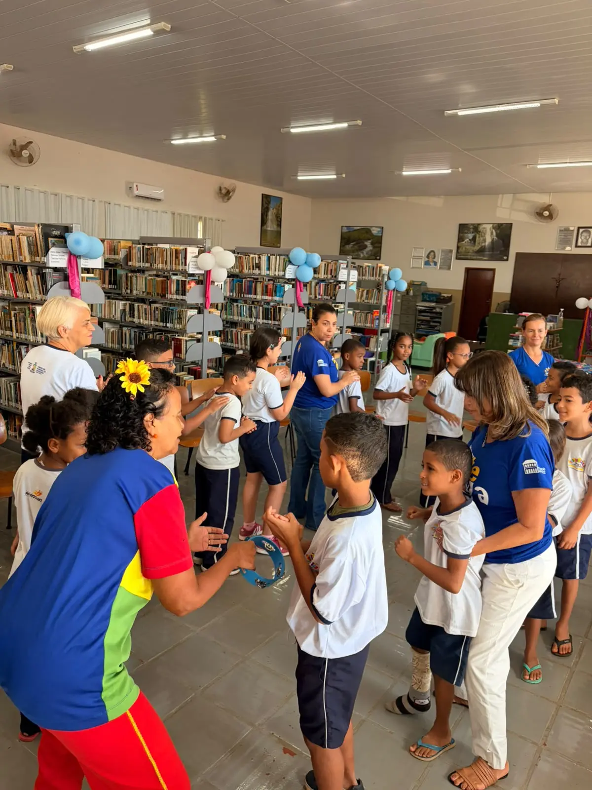 Alunos de escola municipal em brincadeira de roda com pandeiro entre as estantes, na Aventura na Biblioteca