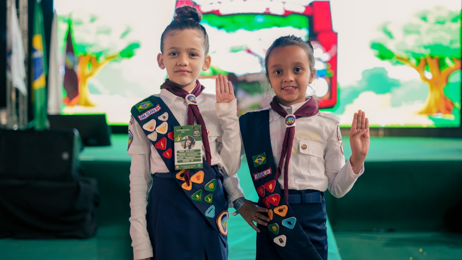 Duas crianças uniformizadas de Aventureiros acenando diante do palco