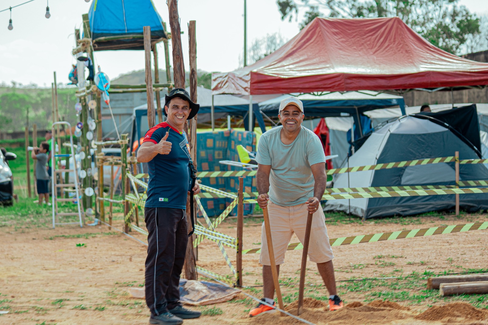 Dois voluntários posam com ferramentas diante de barracas