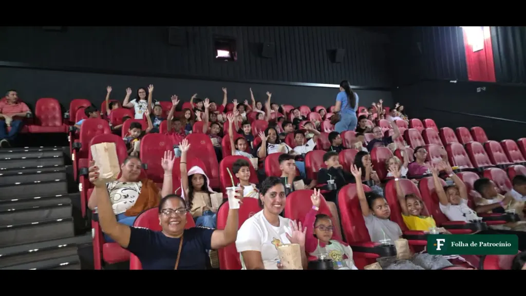 Sala de cinema cheia de crianças com os braços levantados durante o Cine Diversão em Patrocínio