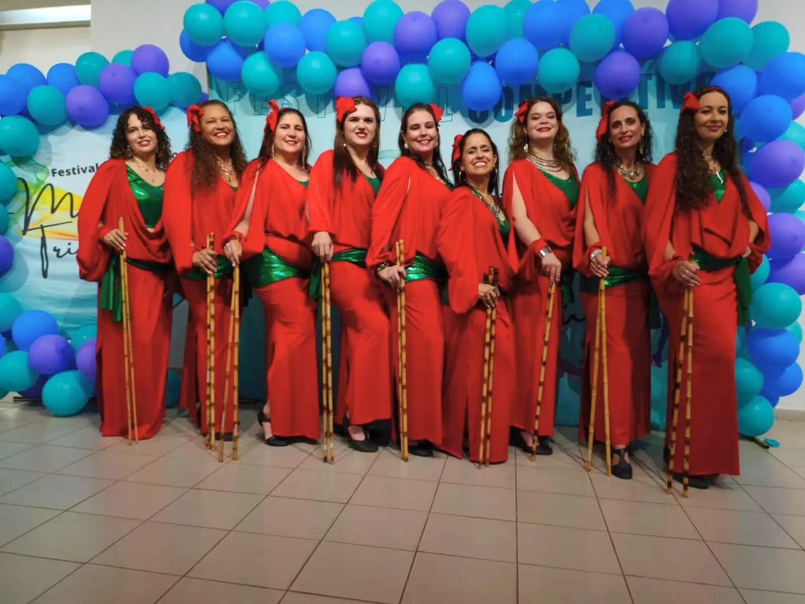 Nove bailarinas de dança do ventre de Patrocínio, de vestido vermelho e faixa verde, seguram bastões de bambu diante do painel do Festival Movimenta Triângulo