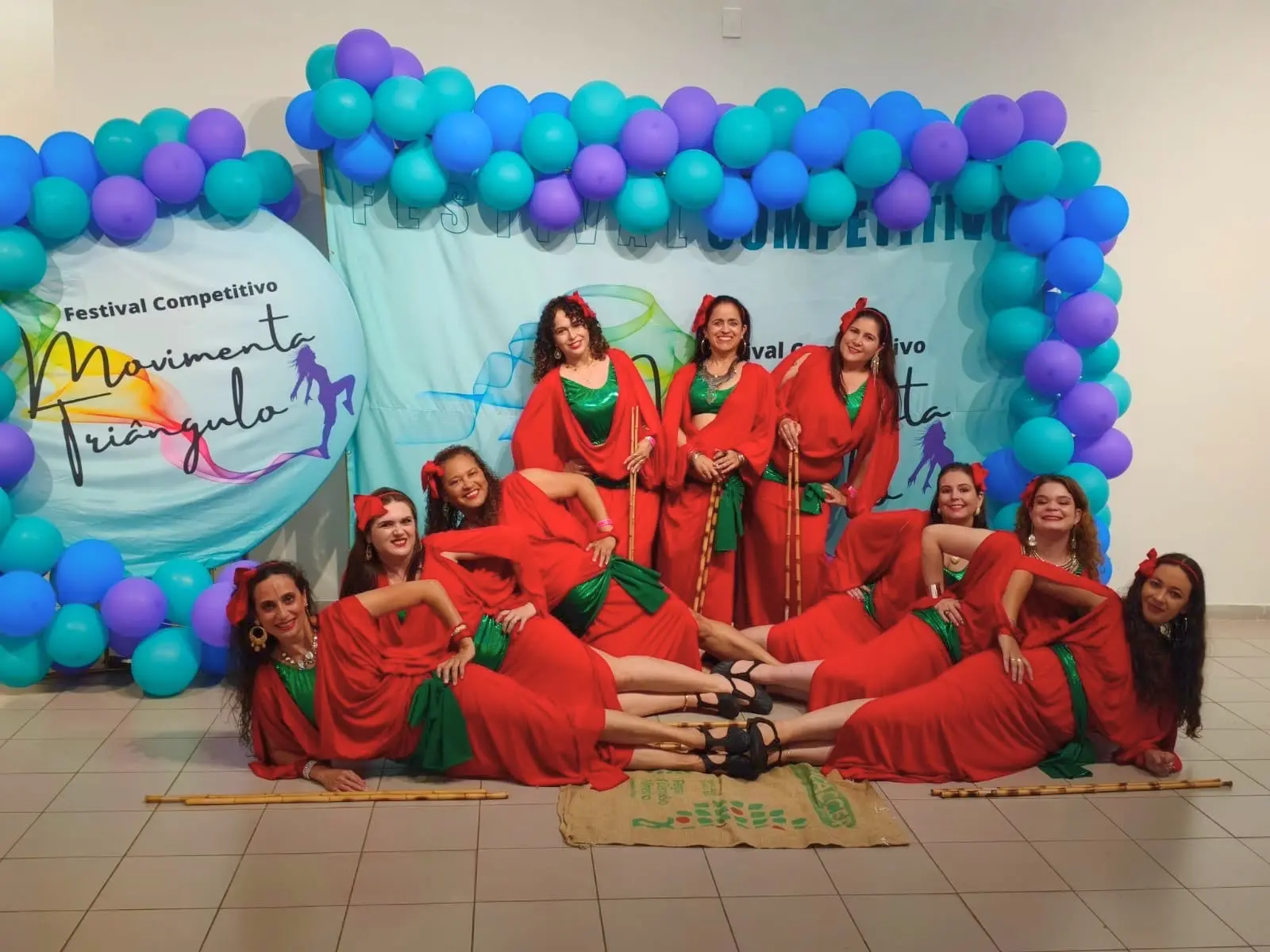 Bailarinas de vermelho posam deitadas e em pé diante do painel do festival, com bastões de bambu e uma saca de café no chão