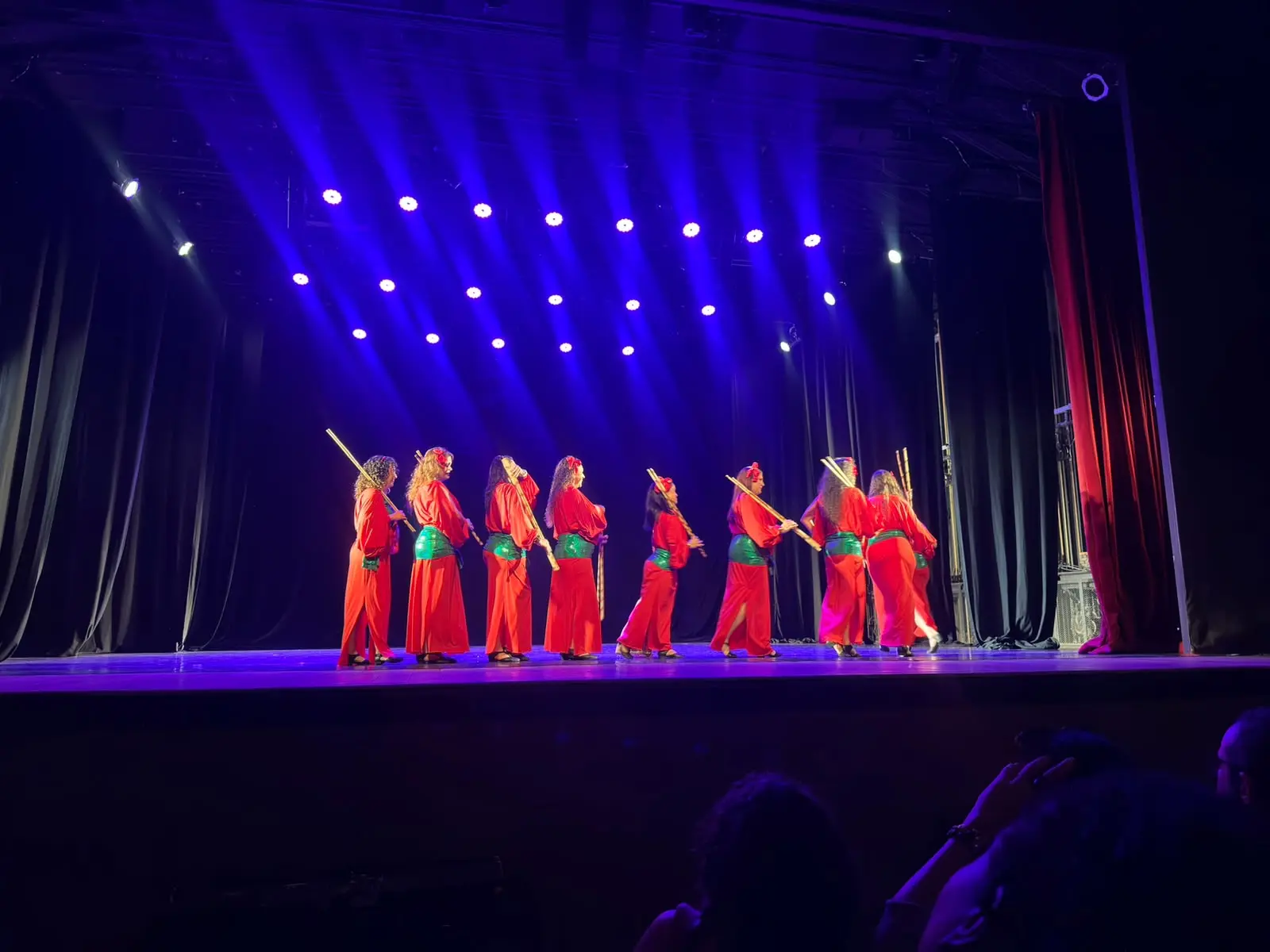 Bailarinas de vermelho em fila no palco, com bastões nas mãos, sob refletores de luz azul