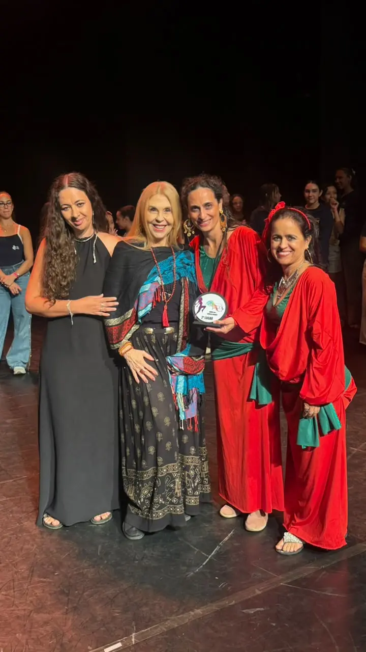 Duas bailarinas de vermelho seguram o troféu no palco, ao lado de duas mulheres de preto