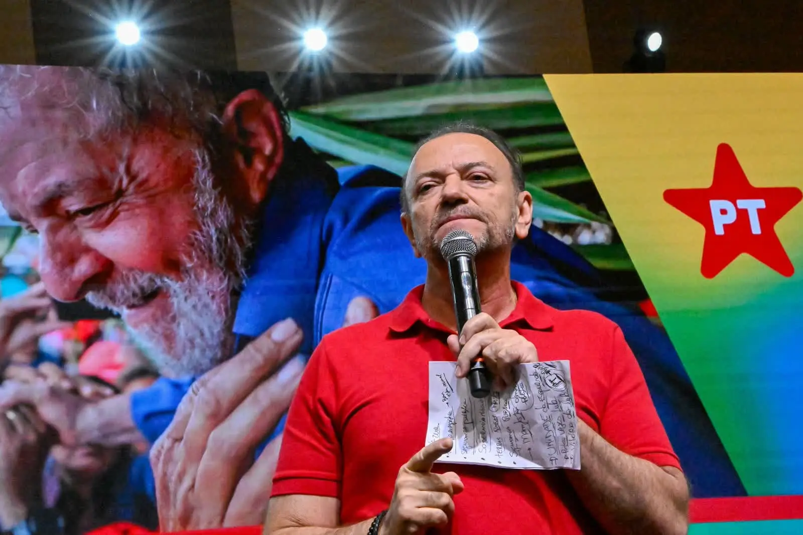 Edinho Silva discursando com o microfone e um papel de anotações diante do telão do PT
