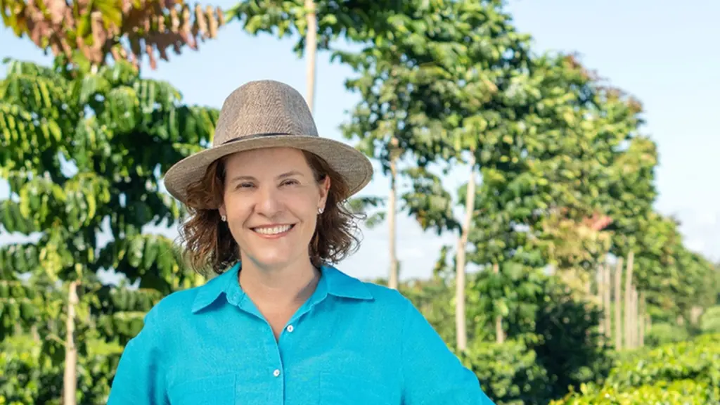 Mariana Velloso Heitor, de chapéu e camisa azul, sorri em meio a uma lavoura de café