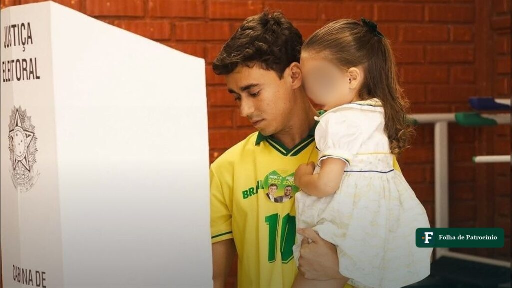 Nikolas Ferreira vota em cabina da Justiça Eleitoral, de camisa do Brasil, com uma criança de rosto desfocado no colo