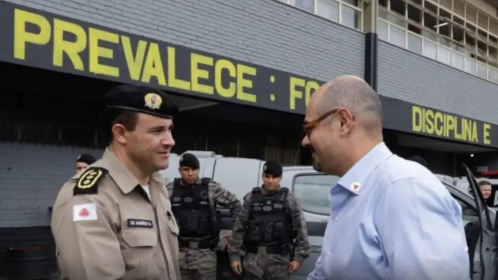 Governador Mateus Simões cumprimenta oficial da Polícia Militar em frente a um quartel, com policiais ao fundo