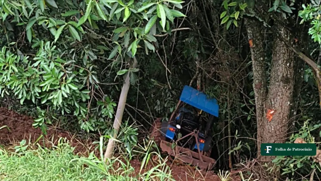 Trator azul tombado contra árvores em encosta na zona rural, local do acidente com trator em Monte Carmelo