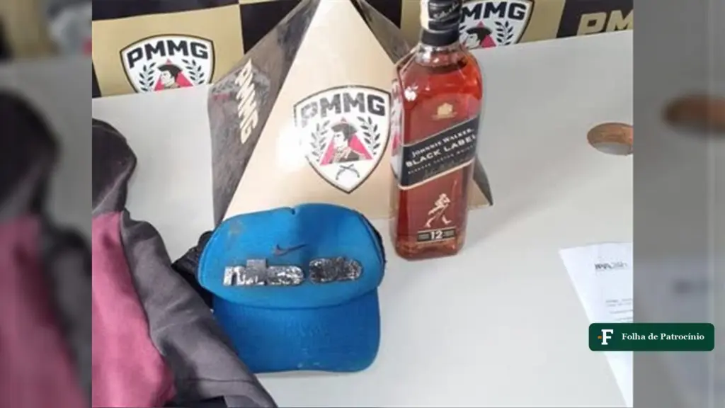 Garrafa de uísque furtado, boné e caixa apreendidos pela PM sobre uma mesa com o brasão da PMMG ao fundo