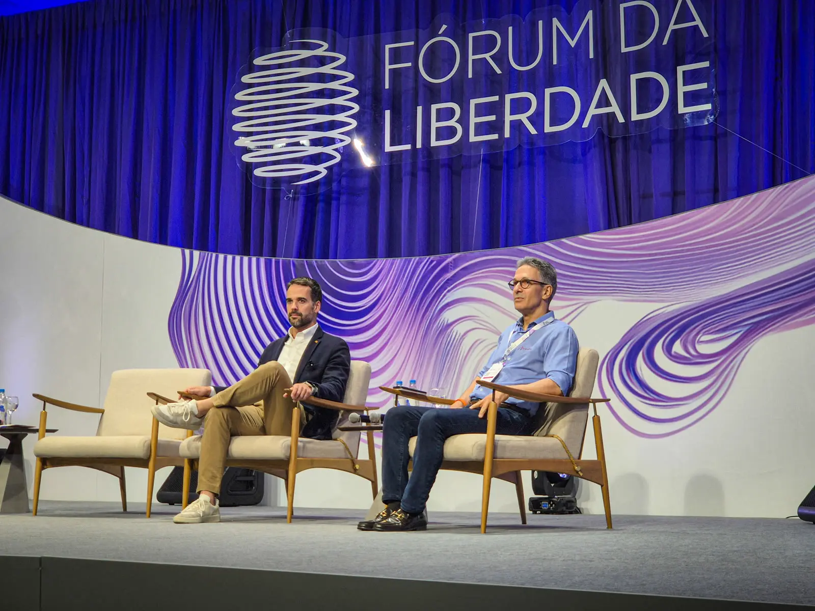 Romeu Zema sentado no palco do Fórum da Liberdade, foto de arquivo sobre Zema e o celular na cabine